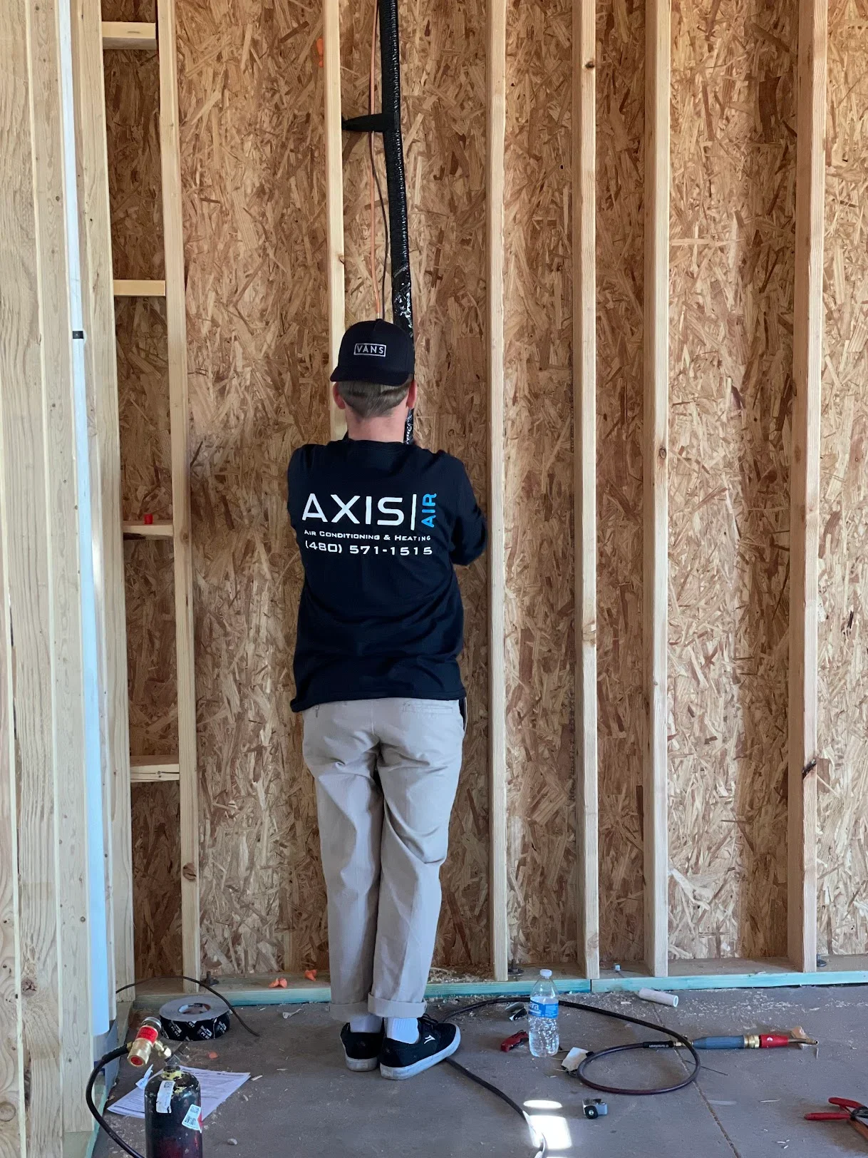 HVAC technician installing ductwork in residential wall framing with OSB sheathing and wood studs